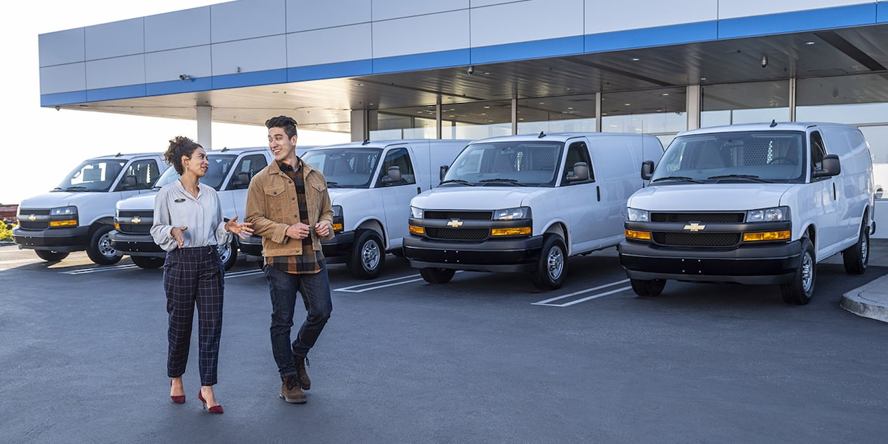 Un vendeur et un client marchent devant plusieurs fourgonnettes Chevrolet blanches stationnées dans une concession.